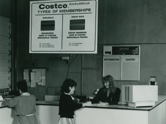 Clientas en el mostrador de un Costco de los primeros años bajo un cartel con los tipos de membresía Wholesale y Group Gold
