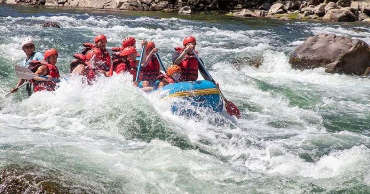 Un grupo con chalecos y cascos rojos rema en una balsa azul por los rápidos de un río entre rocas y árboles