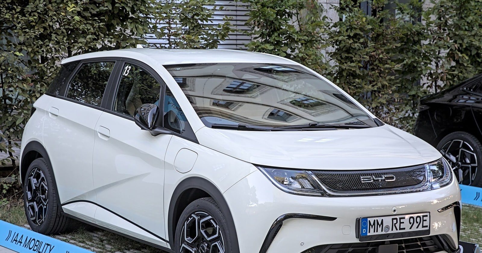 Un BYD Dolphin blanco estacionado sobre una plataforma de exhibición en un salón del automóvil, con árboles de fondo