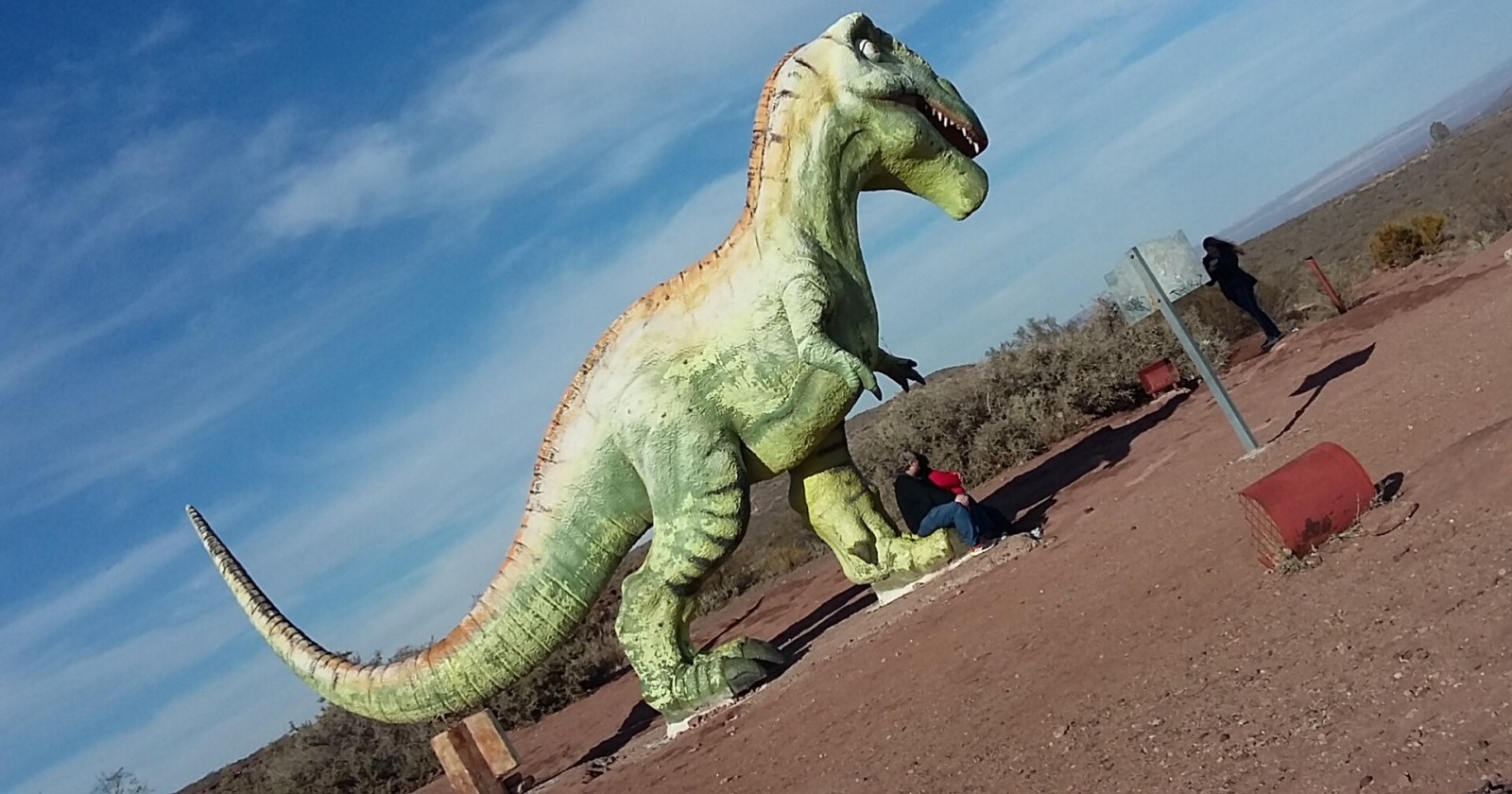 Escultura de dinosaurio carnívoro sobre terreno rojizo en el acceso a Villa El Chocón, con visitantes al pie