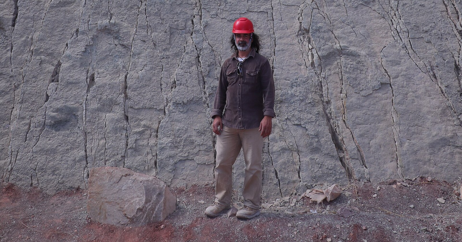 Un hombre con casco rojo parado al pie de una pared de roca inclinada cubierta de huellas de dinosaurio.