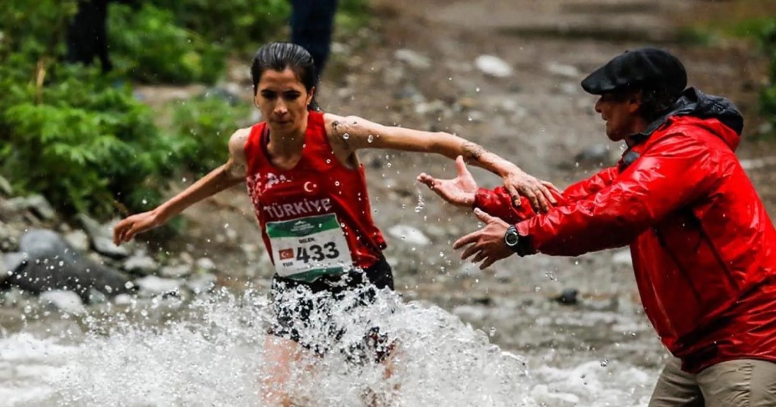 Una corredora con camiseta de Turquía cruza un arroyo mientras un hombre de campera roja le tiende la mano