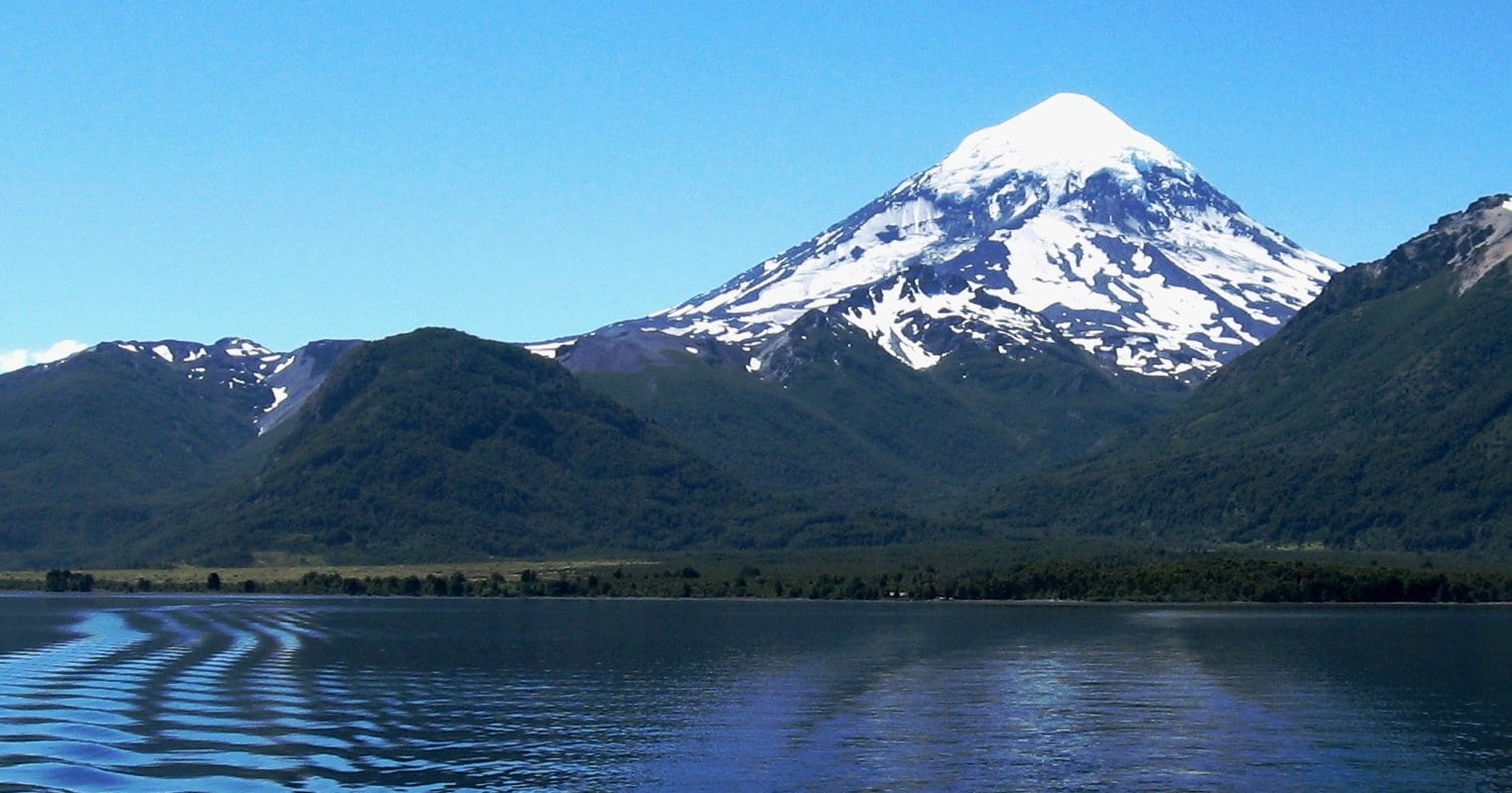 Volcán Lanín nevado sobre bosques y el lago Huechulafquen, con la estela de una embarcación en el agua en primer plano