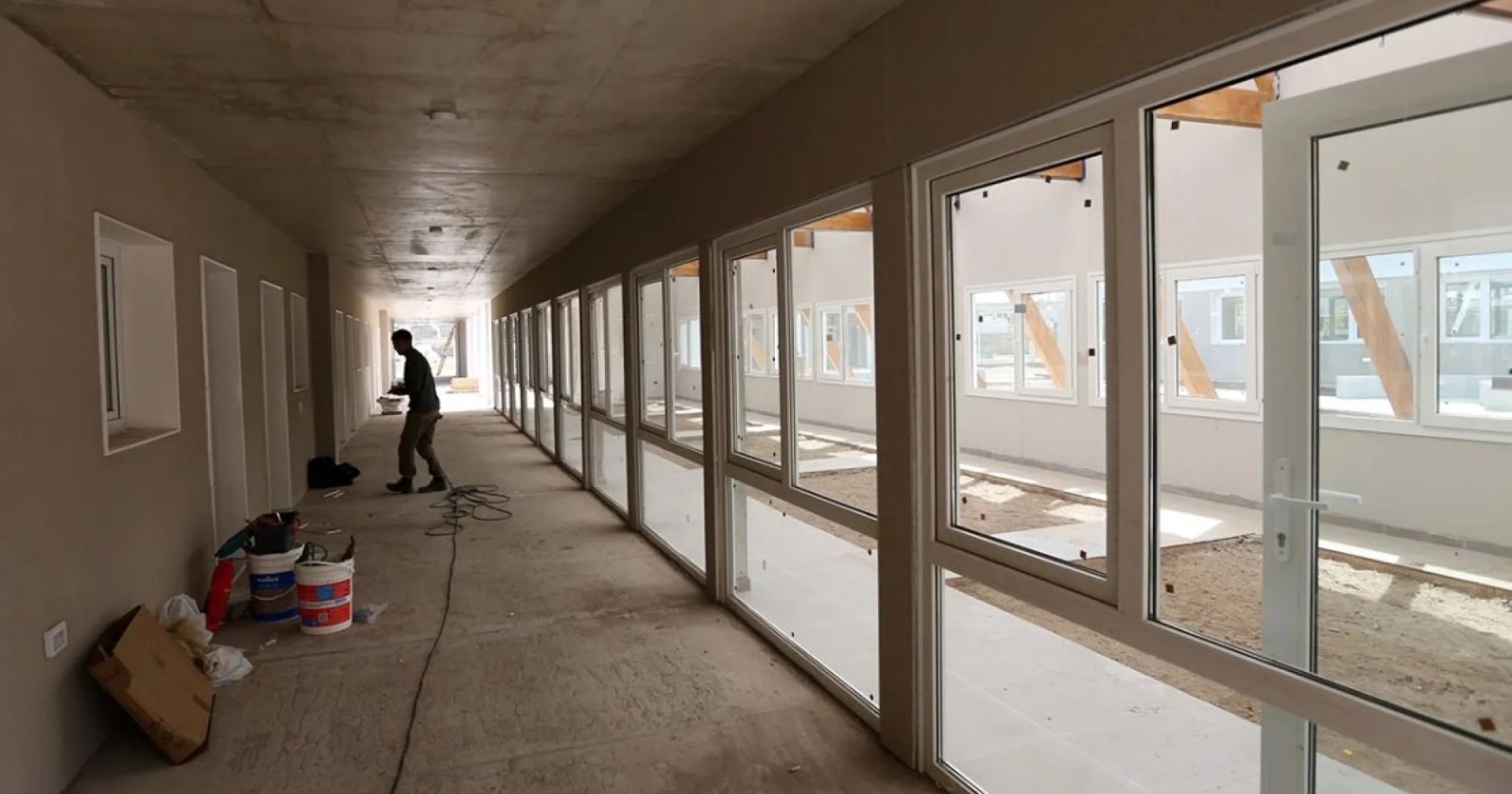 Pasillo de un edificio escolar en obra, con ventanales nuevos, baldes de pintura y un operario al fondo