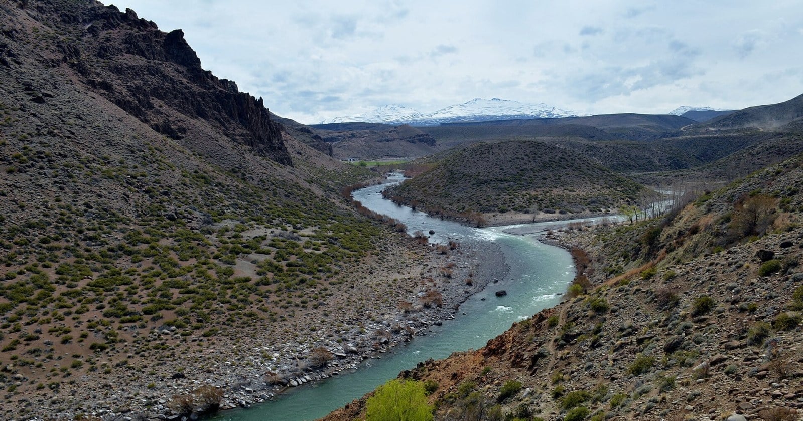 Río Varvarco de agua verde serpentea entre cerros áridos; al fondo, cordillera nevada bajo cielo nublado